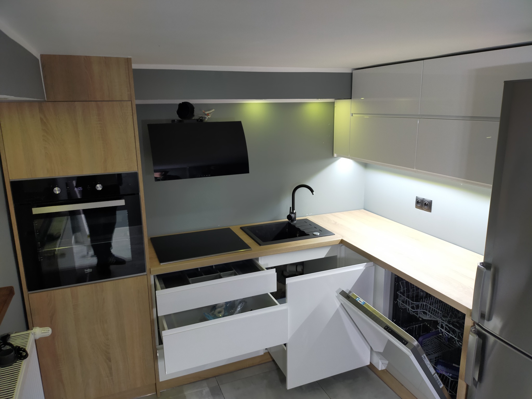 Nowoczesna kuchnia z jasnymi drewnianymi frontami, otwartymi szufladami z organizerami, czarnym zlewem i płytą, oświetleniem LED pod szafkami wiszącymi i częściowo otwartą zmywarką.