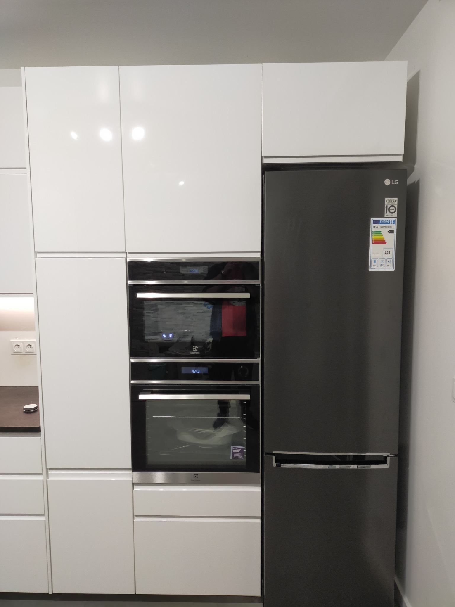 Nowoczesna kuchnia na wymiar z białymi, błyszczącymi frontami, zabudowanym piekarnikiem i mikrofalówką marki Electrolux oraz czarną lodówką LG.