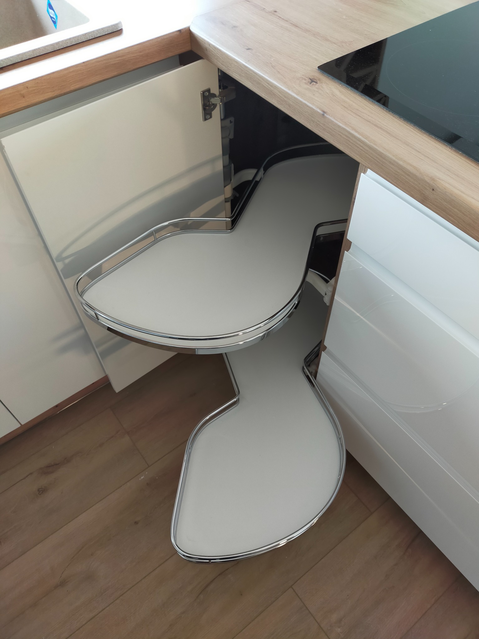 Wysuwany, dwupoziomowy system półek narożnych w szafce kuchennej z białymi frontami i blatem w kolorze jasnego drewna, widoczny na tle drewnianej podłogi.