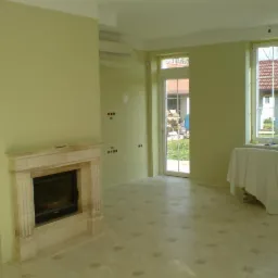 Wnętrze po odświeżeniu, ściany w odcieniu pistacjowej zieleni, marmurowy kominek, zakryte meble, widoczne ślady po gniazdkach elektrycznych.