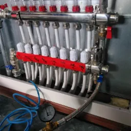 Rozdzielacz ogrzewania podłogowego z rurami, zaworami termostatycznymi i manometrem, widoczne oznaczenia pomieszczeń na czerwonej taśmie.