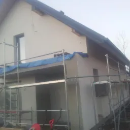Dom jednorodzinny w trakcie wykonywania elewacji, widoczne rusztowania i zabezpieczenia folią. Świeżo otynkowana ściana budynku w kolorze jasnego beżu.