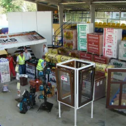 Ekspozycja materiałów budowlanych na targach: dachówki ceramiczne, okna PCV, materiały izolacyjne (styropian, wełna), bloczki silikatowe i narzędzia. Widoczny namiot wystawienniczy firmy Rolteks...