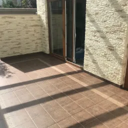 Zadaszony taras z brązowymi płytkami podłogowymi, wykończony dekoracyjnymi płytkami brzegowymi, oraz ścianami wyłożonymi jasną imitacją kamienia, z widocznymi drzwiami tarasowymi.