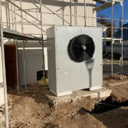 Szara jednostka zewnętrzna z wentylatorem na betonowym fundamencie w Lublinie, otoczona rusztowaniem i wykopem, w tle fragment budynku.