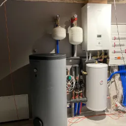 Kompletna instalacja grzewcza w kotłowni z szarym zbiornikiem Trinity, białym zasobnikiem, pompą ciepła Panasonic Aquarea i osprzętem hydraulicznym na tle szarej ściany i surowego stropu.