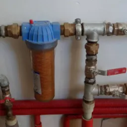 Instalacja wodna z filtrem o zabarwionej zawartości, widoczne złącza i zawory z czerwonymi uchwytami.