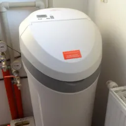 Biały zmiękczacz wody Viessmann z wyświetlaczem i naklejką informacyjną, zainstalowany w pomieszczeniu z rurami i grzejnikiem.