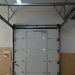 Biała segmentowa brama garażowa z drzwiami wejściowymi, widoczna konstrukcja nośna pod sufitem, w tle okno z widokiem na zewnątrz.