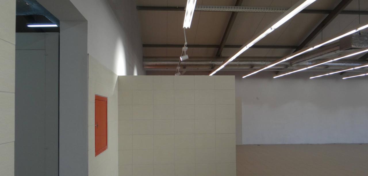 Wnętrze hali przemysłowej z jasną, kafelkową ścianą, pomarańczową skrzynką rewizyjną i rzędami świetlówek pod sufitem.