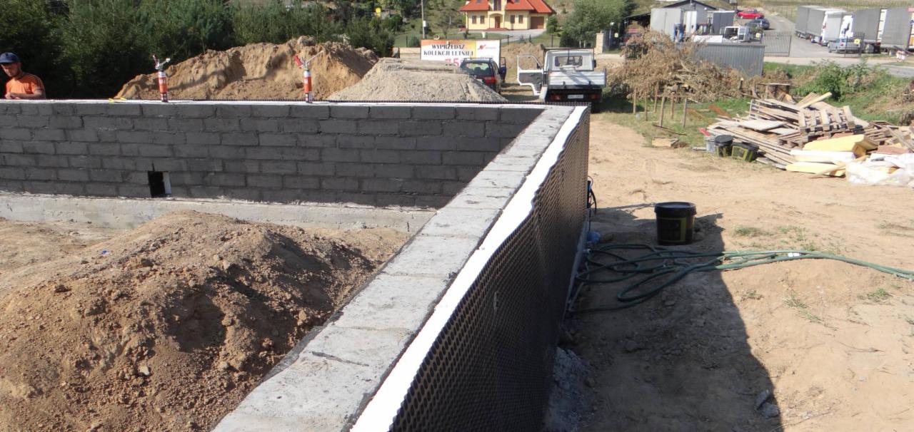 Fundamenty domu z bloczków betonowych z izolacją pionową, widoczne stosy piasku i gruzu, w tle zabudowania i zieleń.