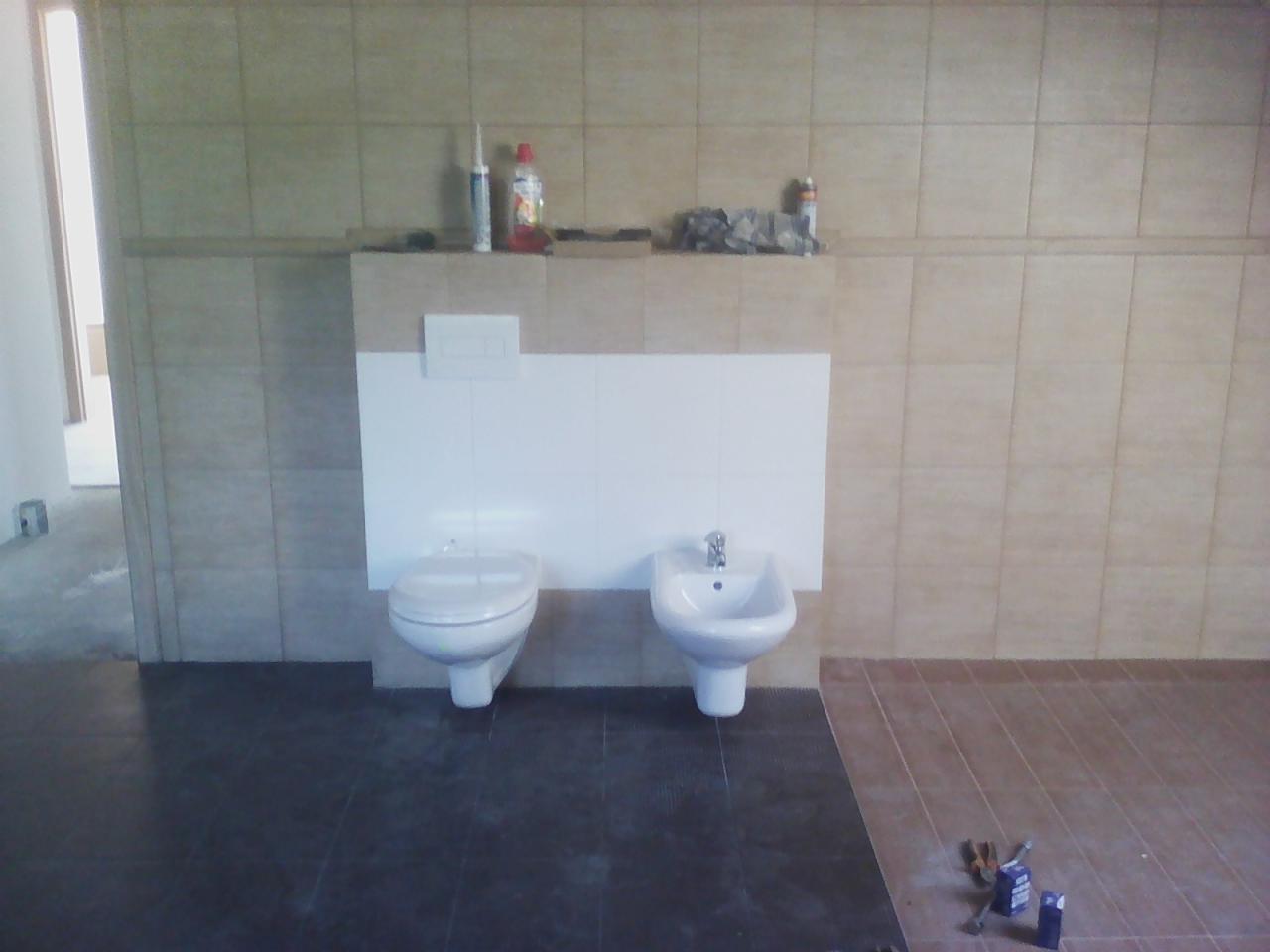 Wnętrze łazienki w trakcie remontu: widoczne białe sanitariaty (toaleta i bidet) zamontowane na tle beżowych płytek, z fragmentem podłogi wyłożonej ciemnymi płytkami oraz narzędzia i opakowania po...