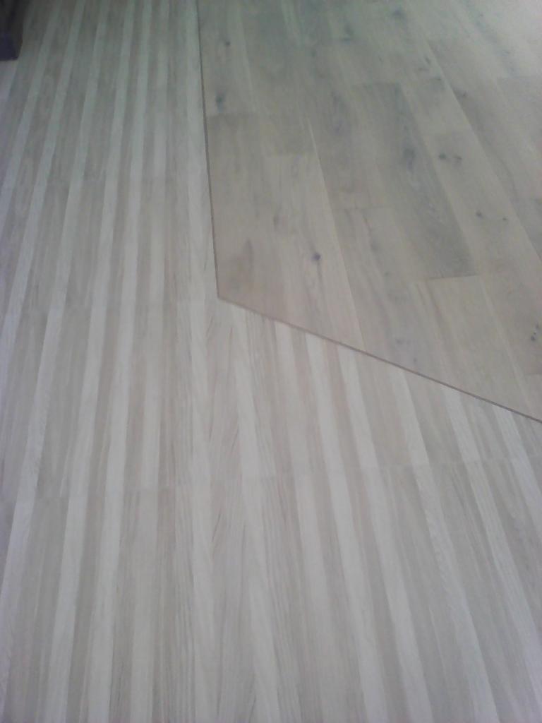 Fragment podłogi z paneli w odcieniach szarości, z widocznym łączeniem dwóch różnych partii paneli o nieco innym odcieniu i wzorze słojów.