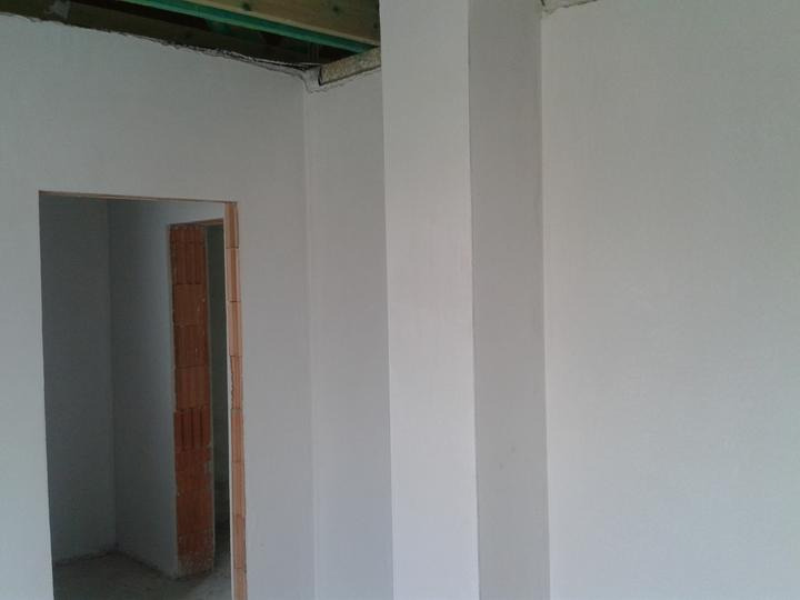 Wnętrze w trakcie prac wykończeniowych: ściany pokryte jasną, świeżą warstwą, widoczny surowy otwór drzwiowy z ceglaną obudową i fragment drewnianej konstrukcji stropu.