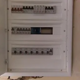 Otwarta skrzynka bezpiecznikowa marki ETI, zamontowana na pomalowanej ścianie z widocznymi uszkodzeniami tynku.