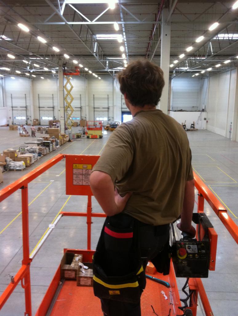 Mężczyzna w uprzęży na podnośniku koszowym, z narzędziami przy pasie, kontroluje montaż oświetlenia w hali magazynowej.