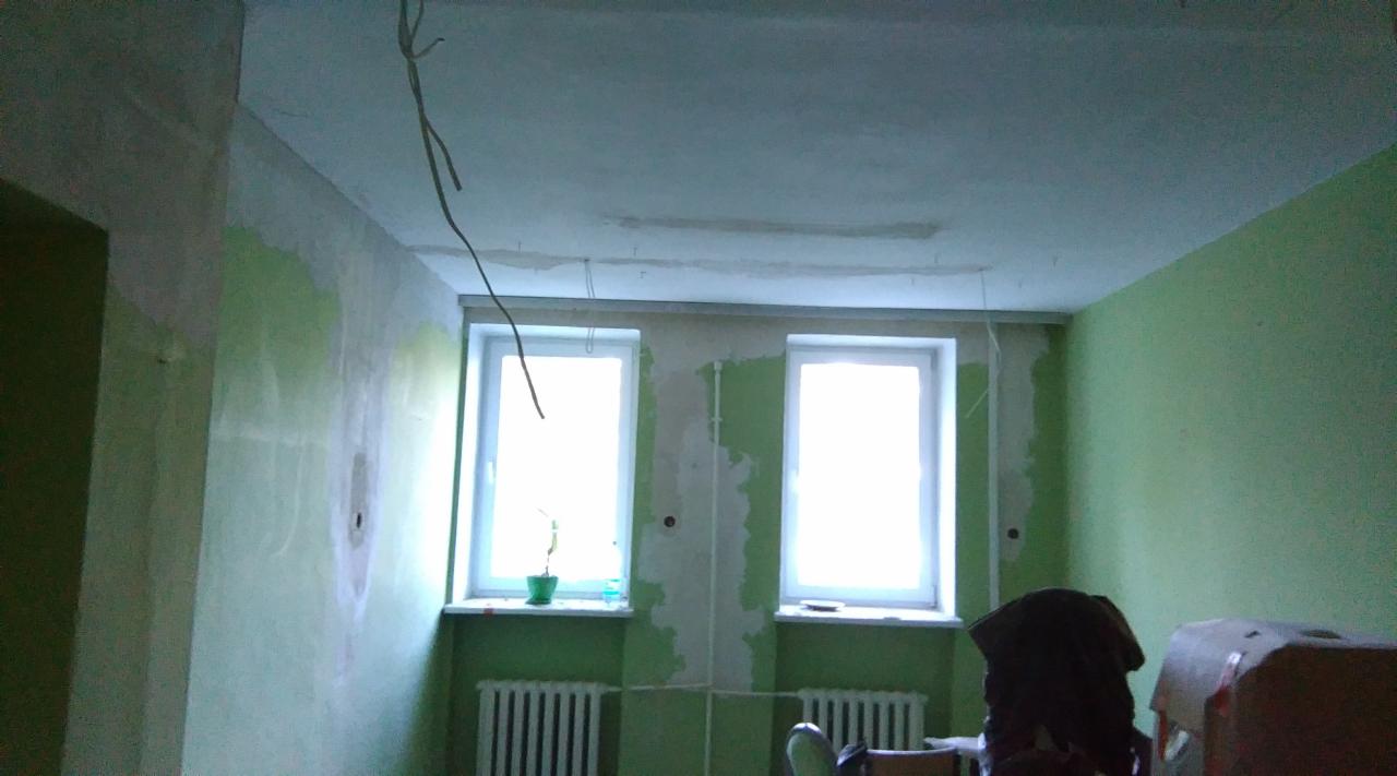 Pokój w trakcie remontu: ściany częściowo pomalowane na zielono, z widocznymi śladami szpachlowania, dwa okna z grzejnikami pod spodem, wystające przewody elektryczne z sufitu, widoczne profile...