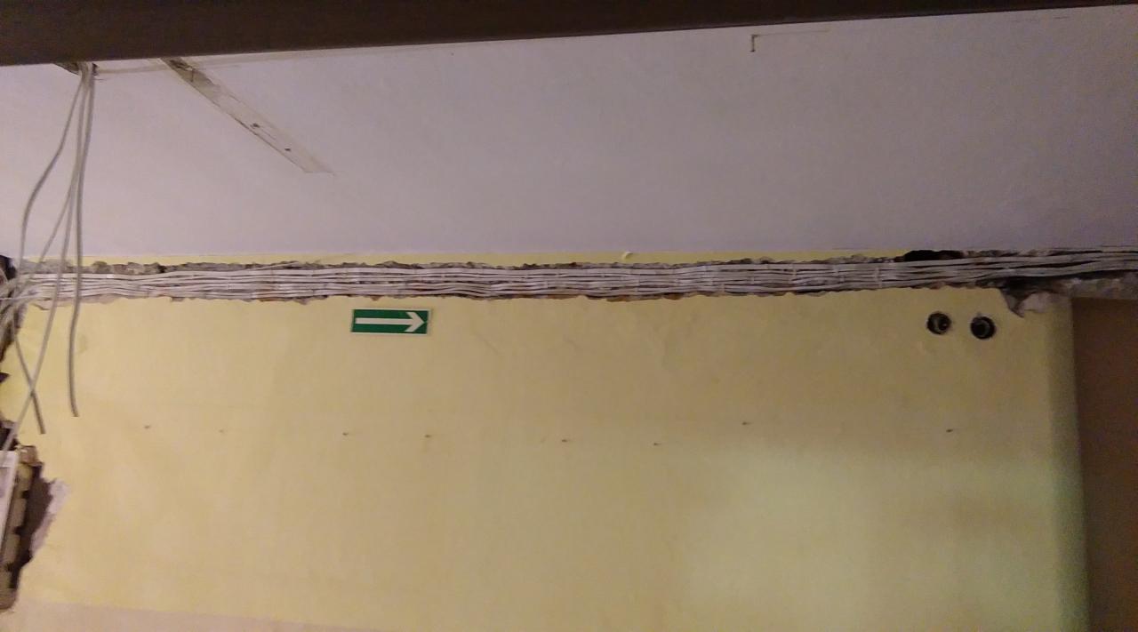 Ściana z żółtą farbą, bruzda z ułożonymi kablami elektrycznymi, wystające przewody, znak ewakuacyjny z zieloną strzałką w prawo, dwa wywiercone otwory na gniazdka.