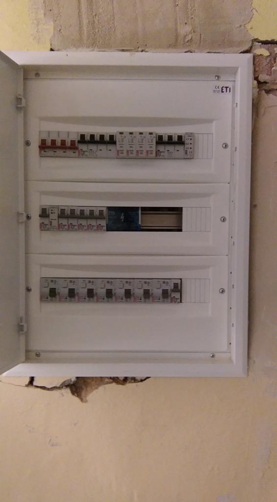 Otwarta skrzynka bezpiecznikowa marki ETI, zamontowana na pomalowanej ścianie z widocznymi uszkodzeniami tynku.