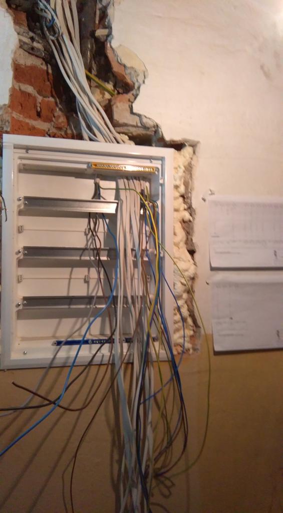 Otwarta skrzynka rozdzielcza z licznymi przewodami wychodzącymi, zamontowana w ścianie z widocznymi śladami skuwania i pianką montażową, na tle fragmentu muru z cegły i dwóch kartek z schematami.