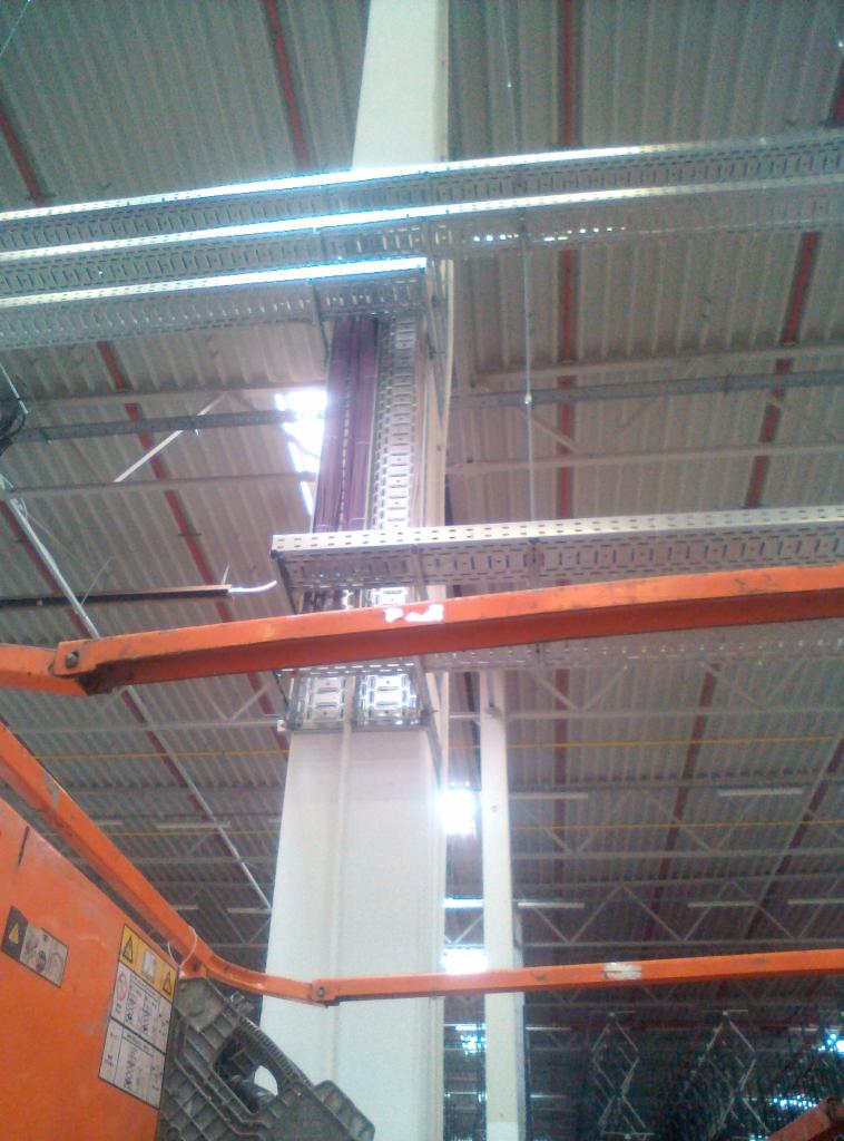 Widok z podnośnika na koryta kablowe i pion kablowy w hali przemysłowej, widoczne okablowanie i konstrukcja dachu.