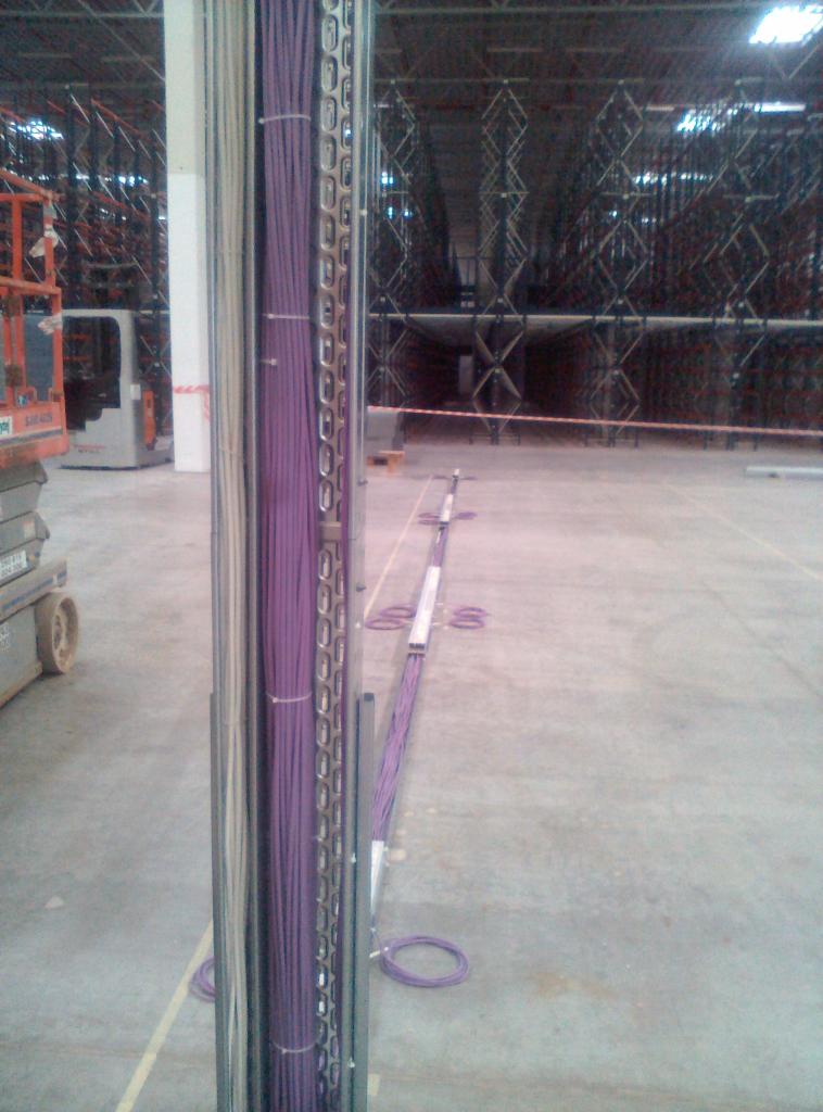 Pionowy korytarz kablowy z fioletowymi przewodami w metalowej drabince, widok w głąb hali magazynowej z regałami i podnośnikiem.