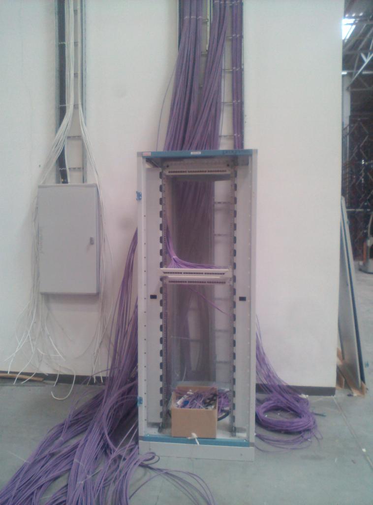 Szafa serwerowa z fioletowymi kablami sieciowymi ułożonymi wewnątrz i na zewnątrz, białe kable na ścianie obok skrzynki elektrycznej, widok na surowy stan wykończenia wnętrza.