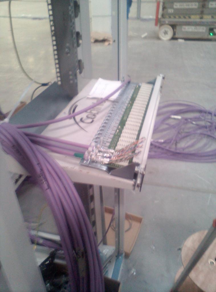 Rozdzielnica telekomunikacyjna z podłączonymi fioletowymi kablami sieciowymi, widoczne złącza i kable w różnych kolorach, na stole warsztatowym z logo firmy.