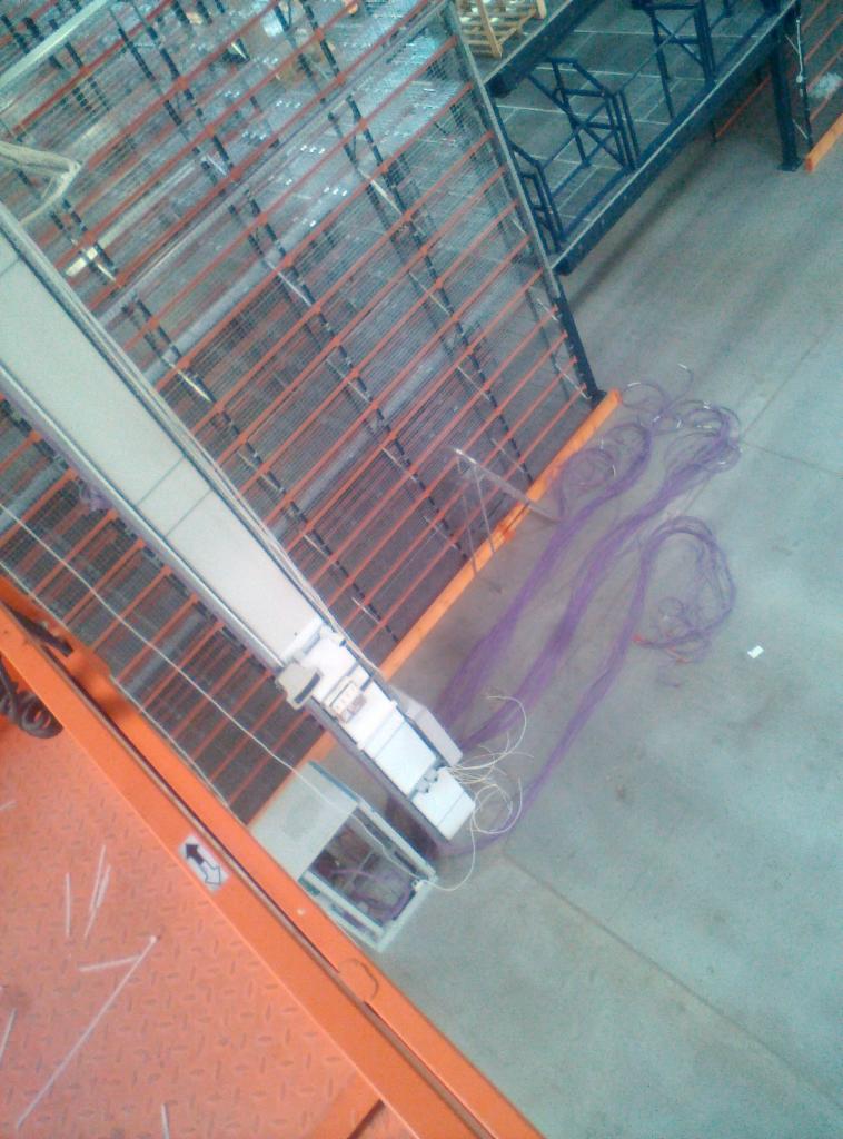 Widok z góry na instalację elektryczną w hali magazynowej, widoczne metalowe regały, kable w fioletowej osłonie ułożone na betonowej podłodze oraz pomarańczowy podnośnik z elementami instalacji.