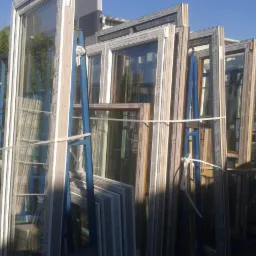 Stos okiennych ram PCV i aluminiowych, zabezpieczonych niebieskimi stojakami i sznurkiem, składowanych na zewnątrz w słoneczny dzień.