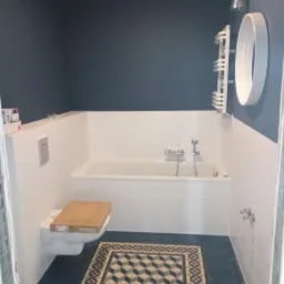 Niebieska łazienka w trakcie remontu: widoczna biała wanna, toaleta z założonym kartonem, grzejnik drabinkowy i wzorzysta podłoga.