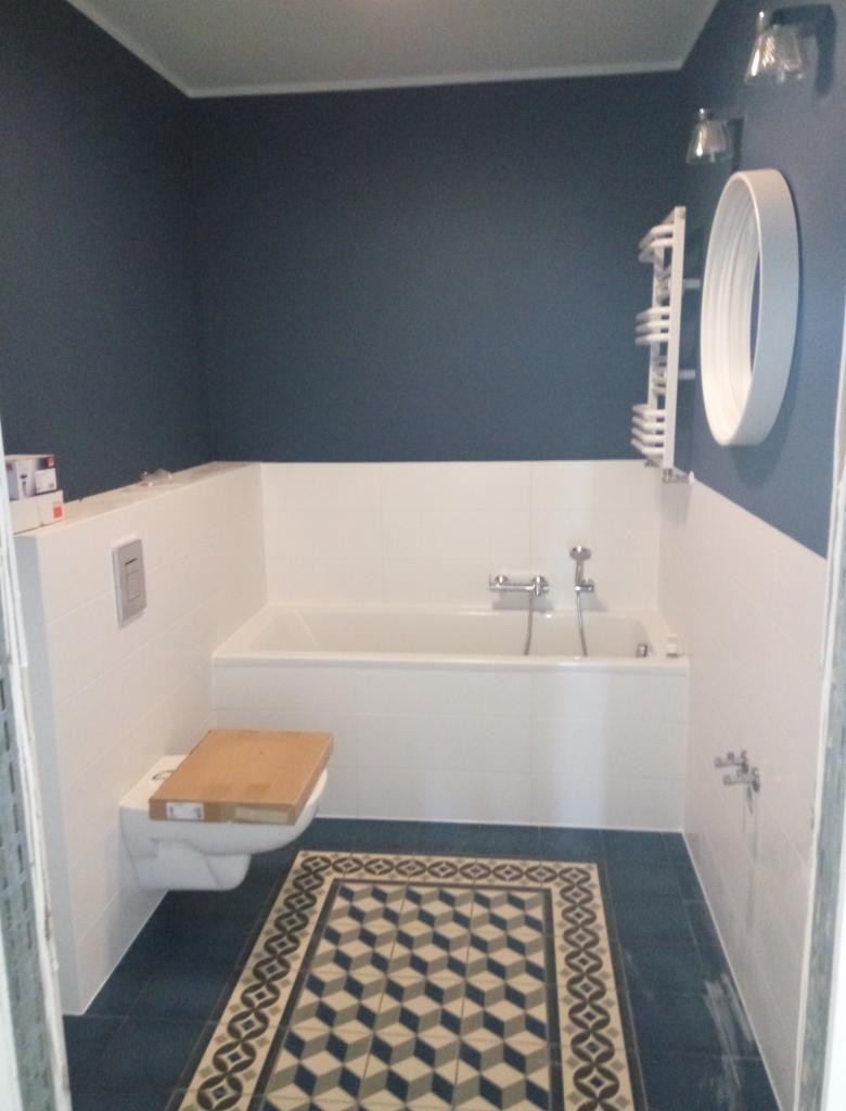 Niebieska łazienka w trakcie remontu: widoczna biała wanna, toaleta z założonym kartonem, grzejnik drabinkowy i wzorzysta podłoga.