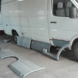 Biały bus w warsztacie z odmontowanymi elementami karoserii, widoczne ślady korozji i przygotowania do naprawy.