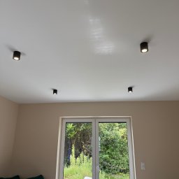 ElektroLuk Łukasz Powała - Wnętrze pokoju z czterema punktowymi lampami sufitowymi w czarnej obudowie, okno balkonowe z widokiem na zieleń ogrodu. Minimalistyczny design.