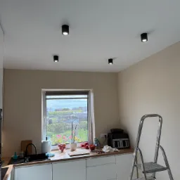 Kuchnia z czterema punktowymi lampami sufitowymi; drabina oparta o ścianę, okno z widokiem na ogród. Minimalistyczny design.