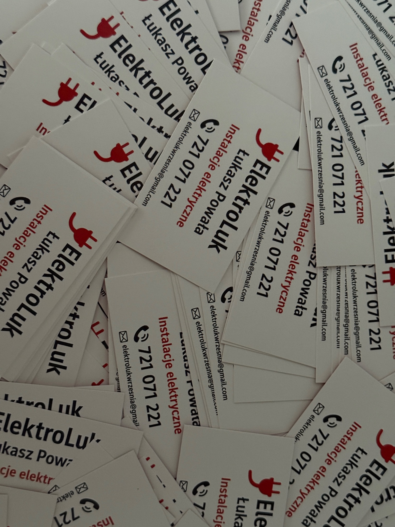 Stos wizytówek elektryka Łukasza Powały z czerwonym symbolem wtyczki i danymi kontaktowymi: numer telefonu i adres e-mail. Widoczne napisy: ElektroLuk, Instalacje elektryczne.