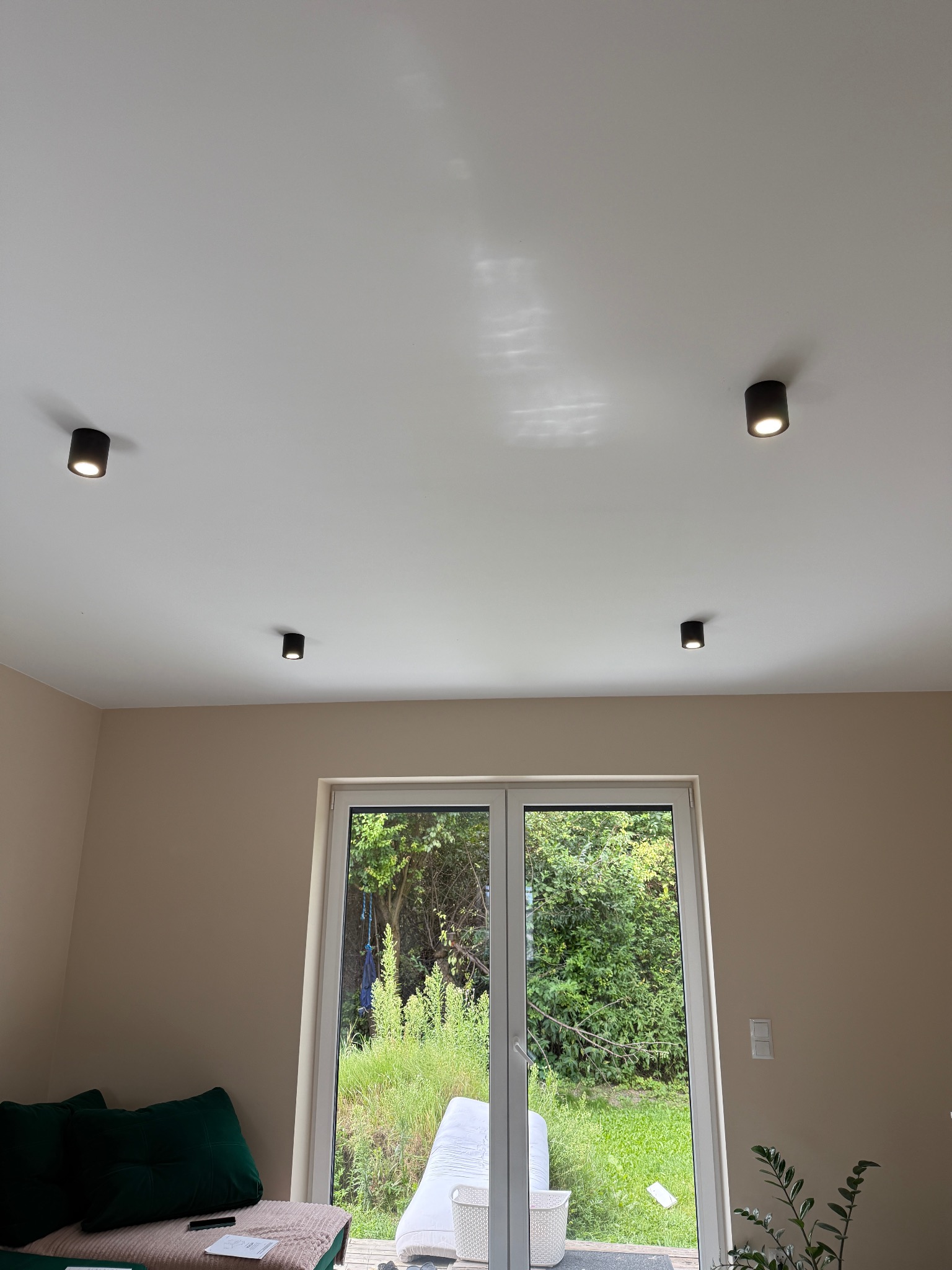 Wnętrze pokoju z czterema punktowymi lampami sufitowymi w czarnej obudowie, okno balkonowe z widokiem na zieleń ogrodu. Minimalistyczny design.