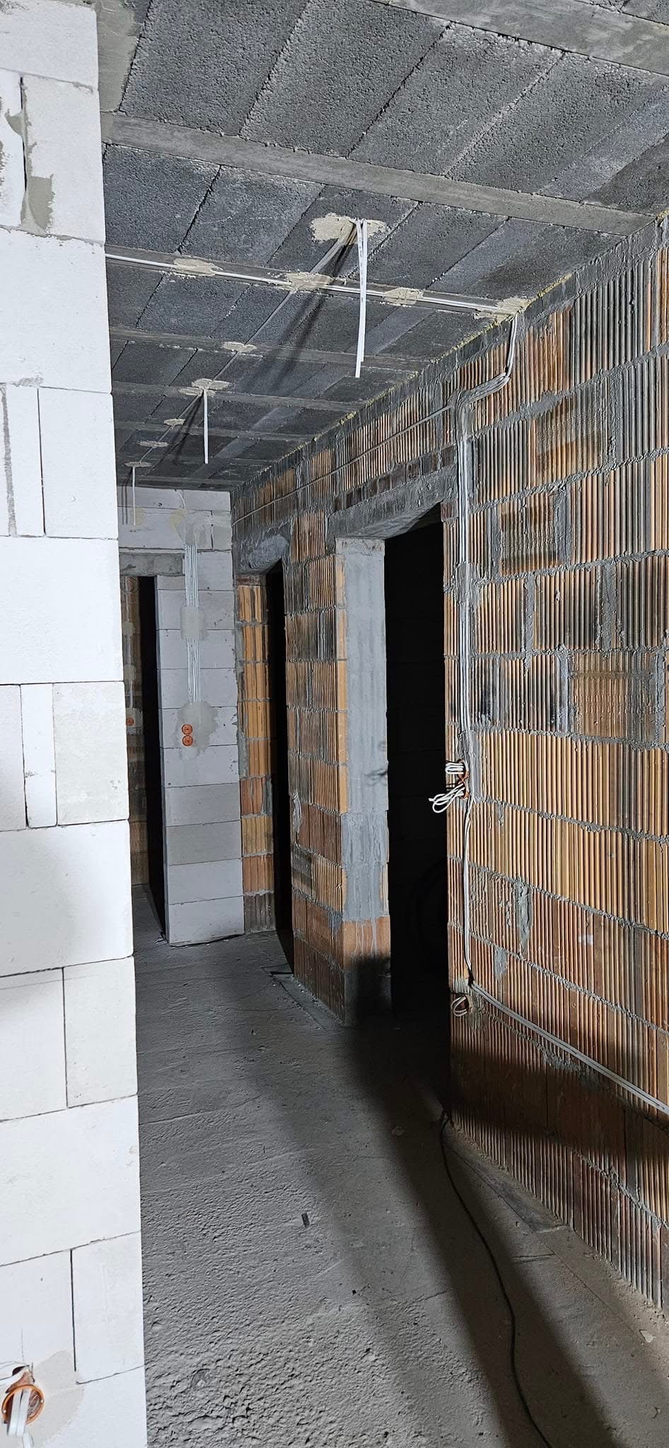 Surowy stan wnętrza: ściany z betonu komórkowego i cegły ceramicznej, wystające przewody elektryczne z sufitu. Widoczne otwory drzwiowe i szara, betonowa podłoga.