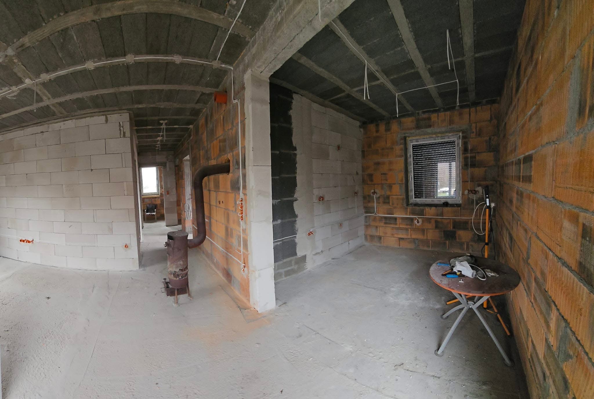 Surowy stan wnętrza z instalacją elektryczną: widoczne kable, puszki, ceglane ściany, betonowa podłoga i sufit. Oświetlenie tymczasowe, piec.