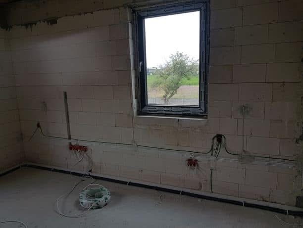 Surowy pokój z widokiem na drzewo przez okno. Instalacja elektryczna w trakcie montażu, widoczne kable i puszki elektryczne na ścianach z betonu komórkowego.