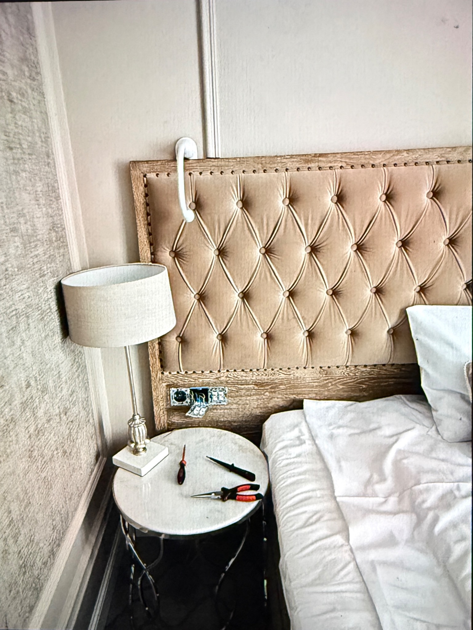 Lampa nocna na stoliku obok łóżka z narzędziami elektrycznymi, widoczna puszka instalacyjna w zagłówku. Przygotowanie do montażu oświetlenia.