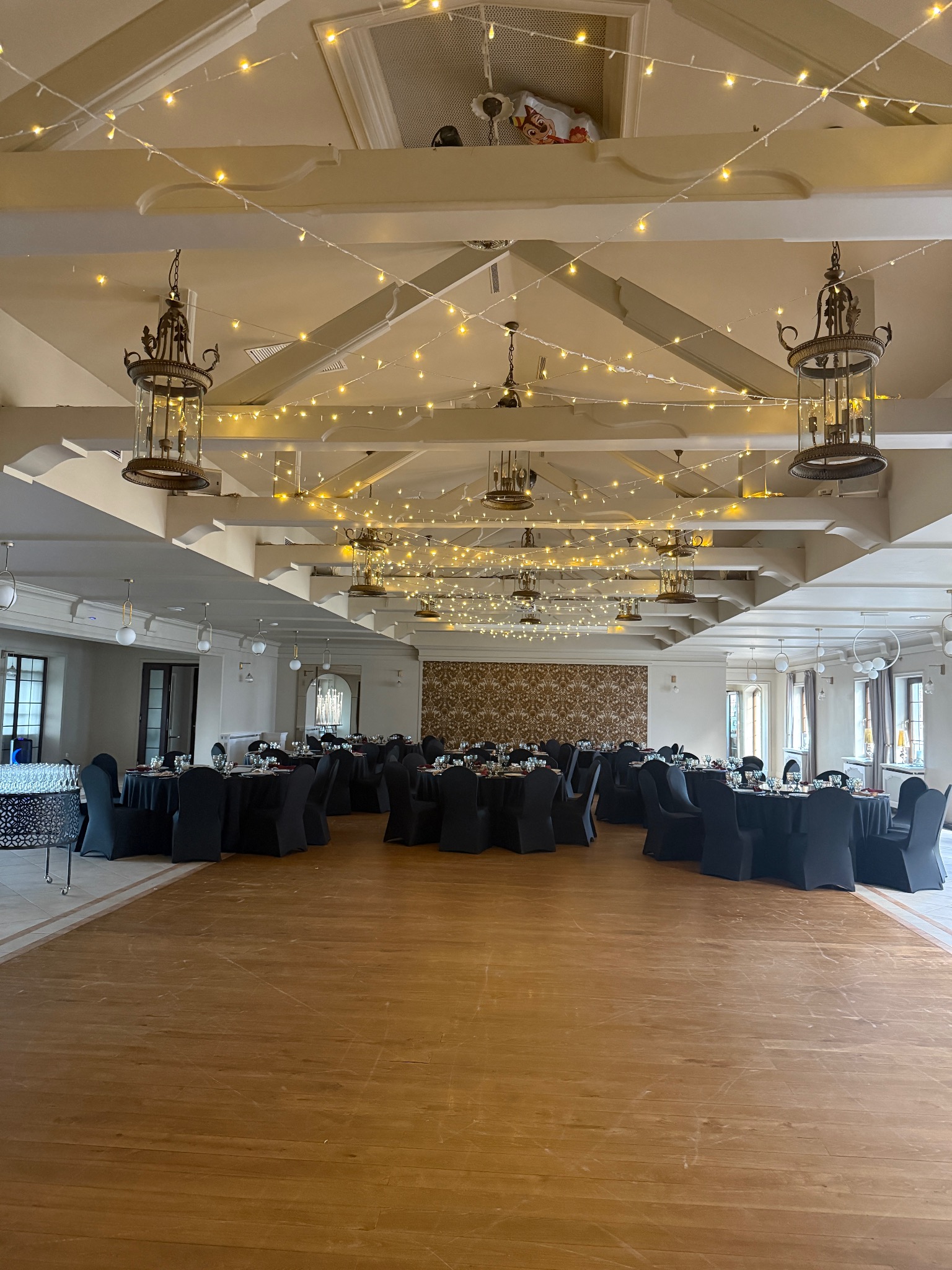 Sala bankietowa z dekoracyjnym oświetleniem: girlandy świetlne i ozdobne żyrandole. Stoły nakryte czarnymi obrusami, parkiet. Przygotowania do uroczystości.