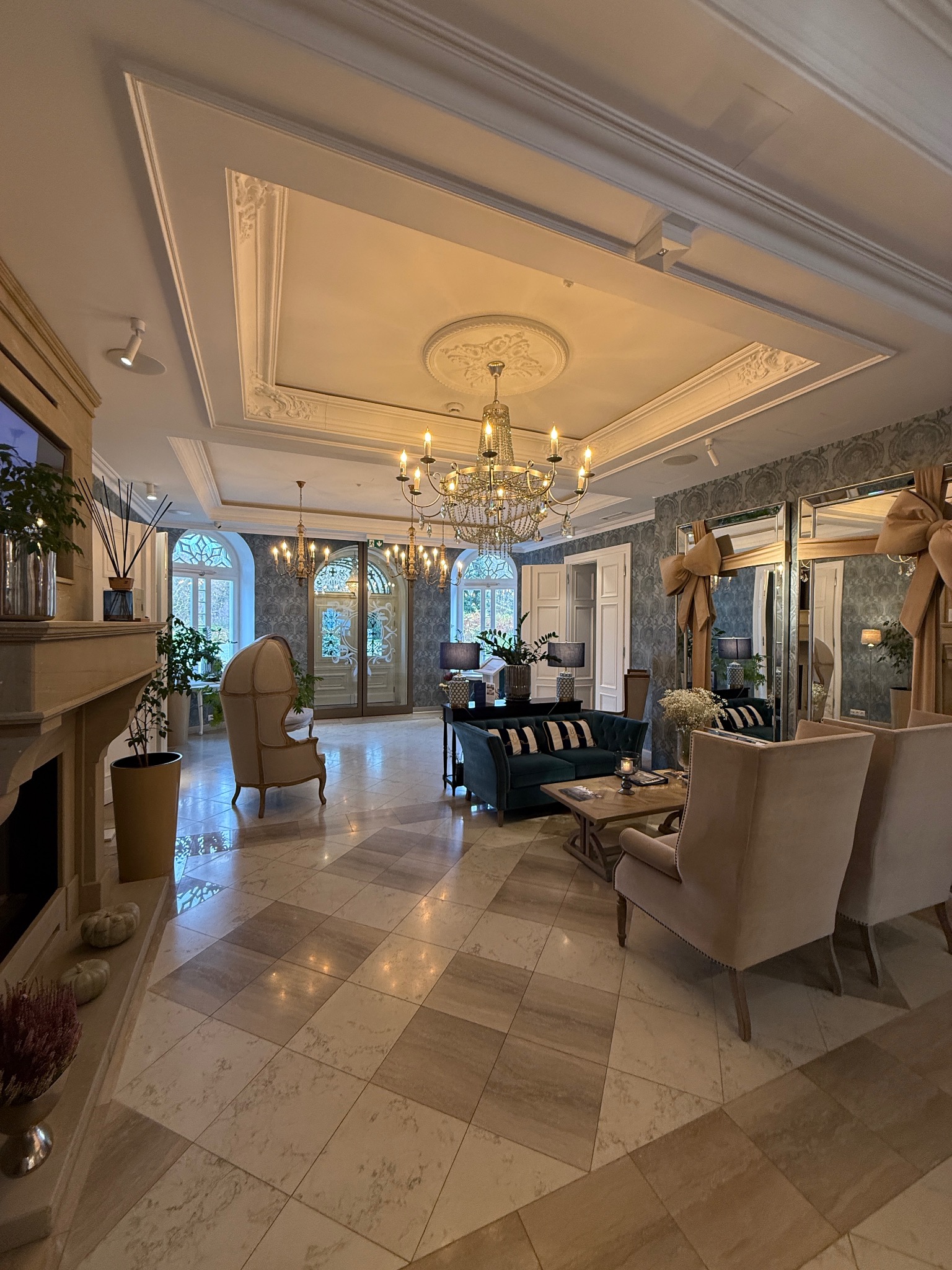 Elegancki salon z żyrandolem, beżowymi fotelami i zieloną sofą. Oświetlenie punktowe w suficie. Parkiet w jasnych odcieniach. Dekoracyjne lustra z kokardami.