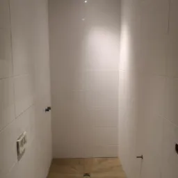 Wąska kabina prysznicowa wyłożona białymi, prostokątnymi płytkami, z drewnopodobną posadzką z odpływem liniowym w kształcie gwiazdy. Widoczne punkty instalacyjne na ścianach i oświetlenie sufitowe.