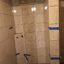 Ściany prysznica wyłożone beżowymi płytkami ceramicznymi, zabezpieczone niebieską taśmą, z widocznymi wystającymi rurami instalacji wodnej przed montażem armatury.