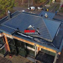 Łącz-dach Paweł Łączyński - Widok z góry na nowoczesny dom z ciemnoszarą dachówką, częściowo pokrytą śniegiem. Widoczne detale architektoniczne i otoczenie budynku. Ujęcie z drona.