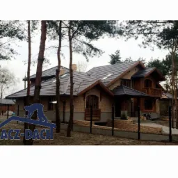 Duży, dwupiętrowy dom z cegły z ciemnoszarym dachem wielospadowym, widoczny z daleka, otoczony drzewami, z logo firmy dekarskiej na pierwszym planie.