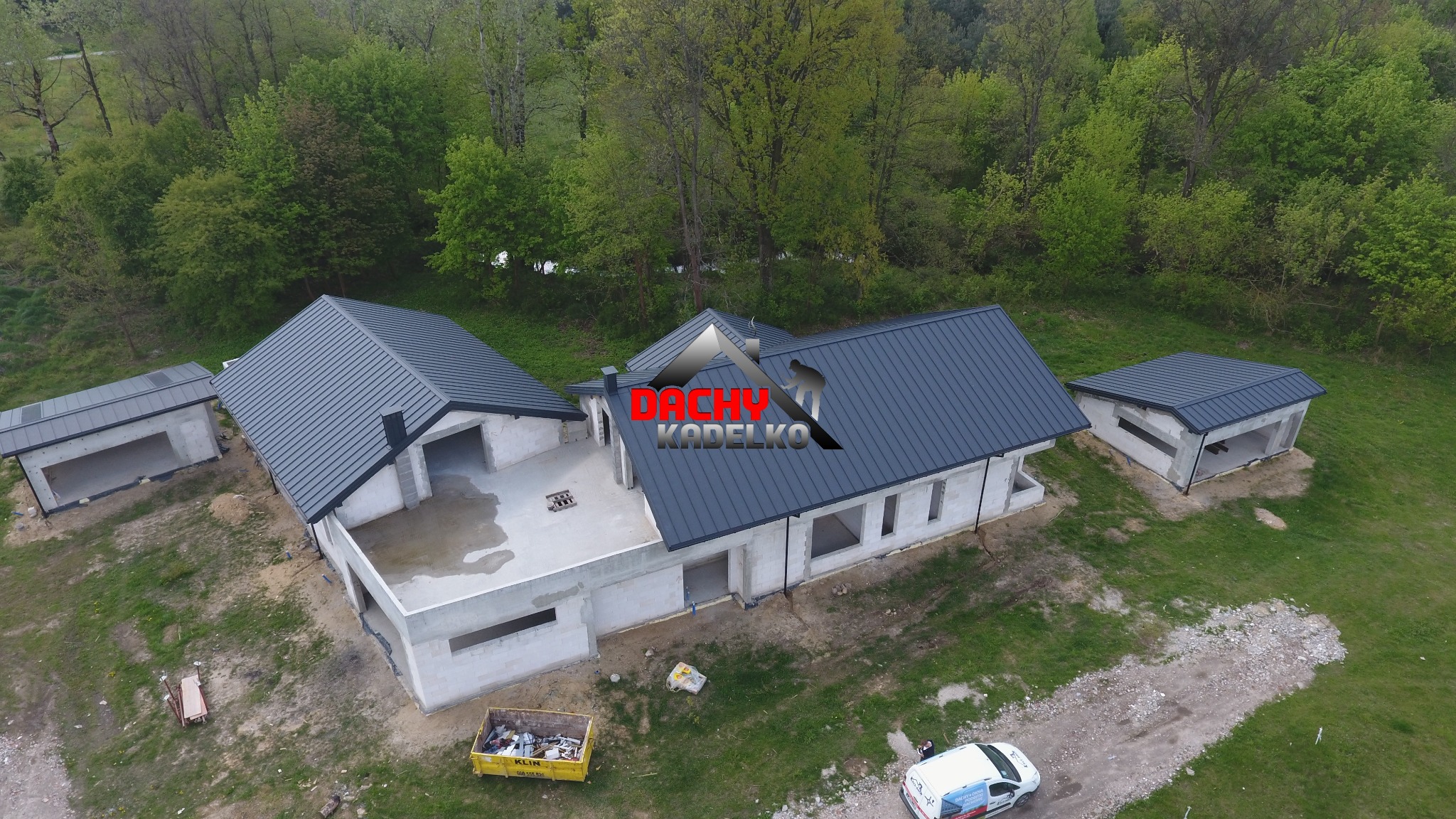 Widok z drona na dom w trakcie budowy z ciemnoszarą blachą trapezową na dachu. W tle las, na pierwszym planie kontener na odpady i samochód dostawczy.