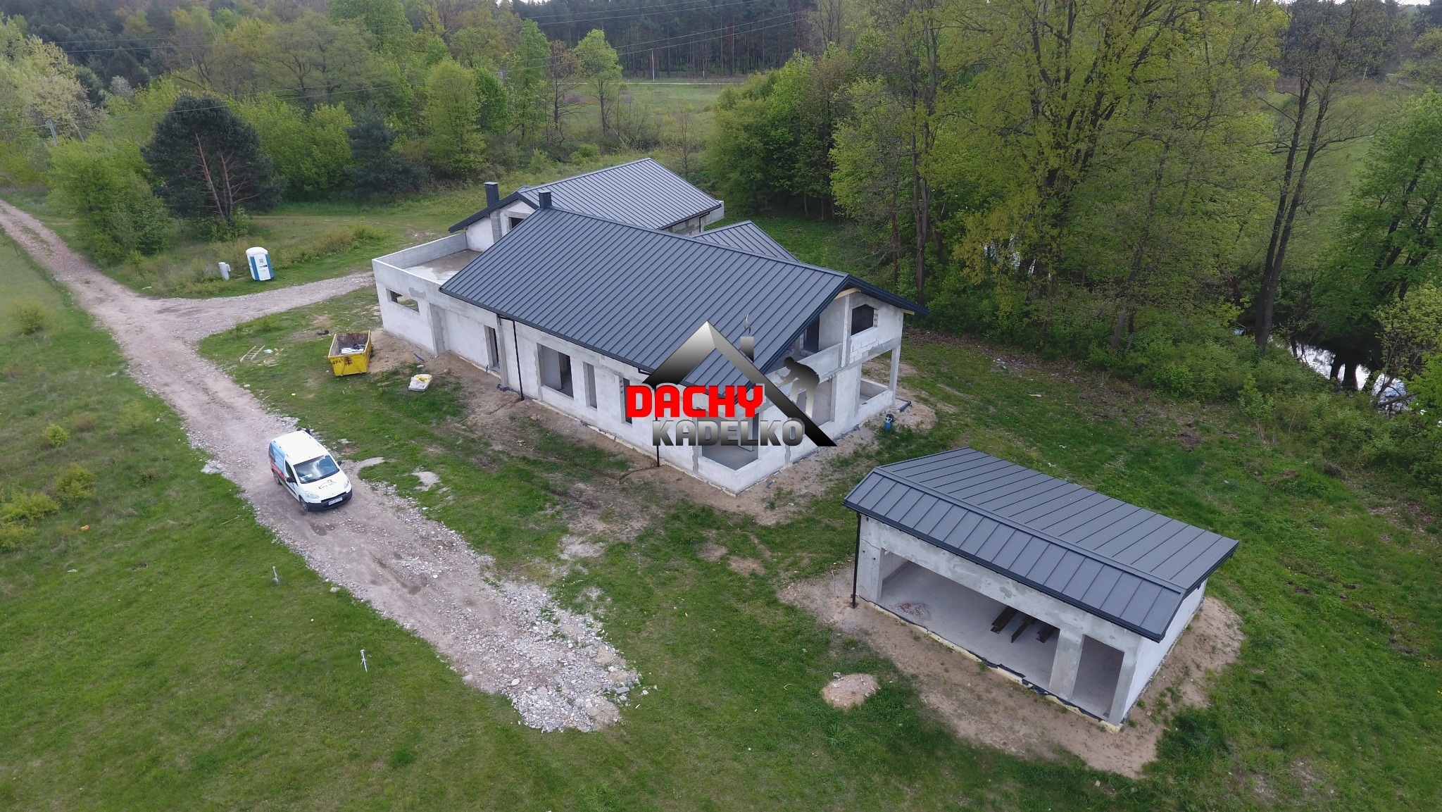 Zdjęcie z drona przedstawia dom w stanie surowym zamkniętym z grafitowym dachem z blachy na tle zieleni. Widoczny także garaż z takim samym dachem i firmowy bus.