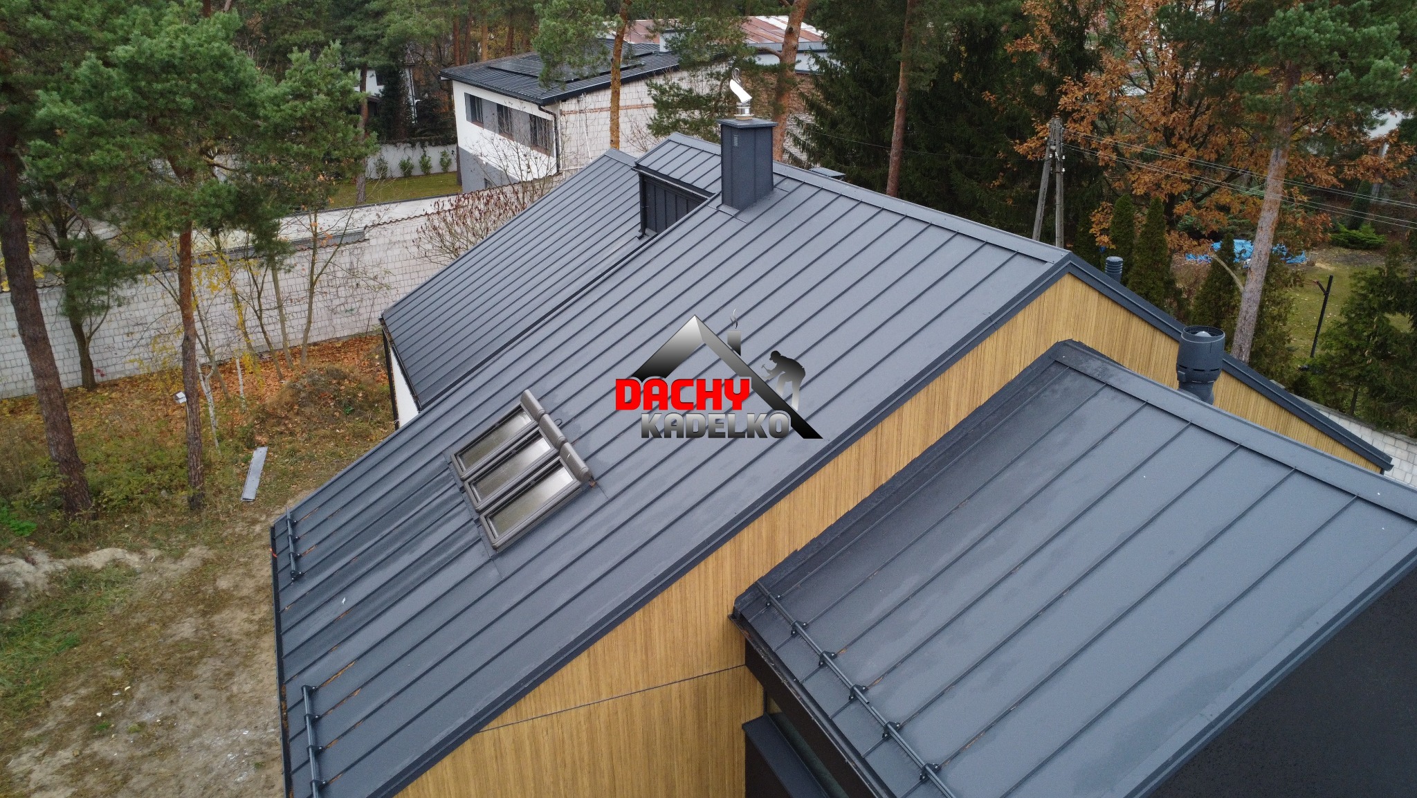 Szary dach z blachy trapezowej z oknami dachowymi na tle drzew. Widok z góry ukazuje estetyczne wykończenie i nowoczesny wygląd pokrycia dachowego.
